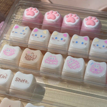 Set of cute cat-themed keycaps in a clear plastic case on a wooden surface.